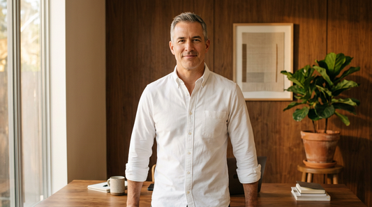 Confident middle-aged man with upright posture standing in a warm-lit home office, wearing a crisp white oxford shirt, illustrating the look of someone using a daily compression shirt for posture support