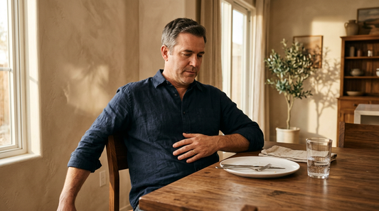 American man, 50, in dark navy linen shirt sitting at a warm walnut dining table after dinner, one hand resting on his stomach — illustrating belly bloat in men over 45